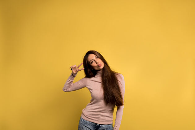 快乐有趣友好的女孩 棕色长发 穿着牛仔裤和粉色衬衫 在黄色的墙上送上一个吻和和平的标志姿态自我积极