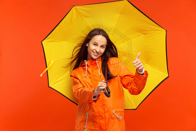 下雨这个快乐微笑的年轻女孩穿着秋天的橘色夹克在摄影棚里摆姿势红色隔离人类积极的情感寒冷天气的概念女性时尚概念外套伞天气