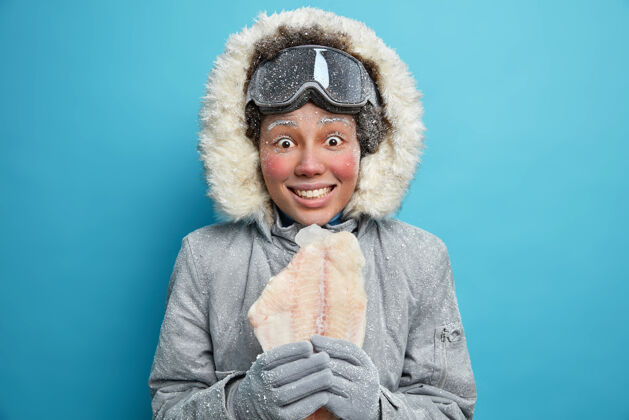 冰北极女探险家在冬天穿着外套去冰上钓鱼 抱着穿着舒适衣服的冻鱼在蓝色的墙上颤抖 在寒冷的日子里为天气变化做准备女人毛皮心情