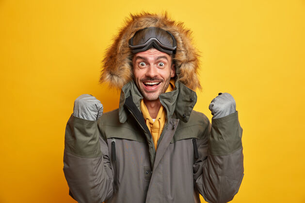 冬季快乐的年轻人欢喜冬天来了举起握紧的拳头戴着手套和带兜帽的暖和夹克帅气积极高兴