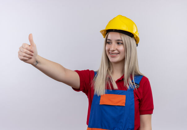 制服微笑的年轻金发工程师建筑女孩在制服和牙套显示拇指在左侧孤立的白色空间秀牙套工程师