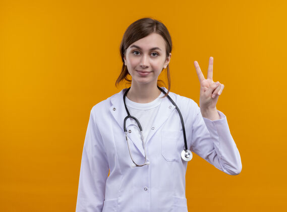 复制高兴的年轻女医生穿着医疗袍和听诊器做和平的标志上孤立的橙色空间与复制空间年轻太空橙色