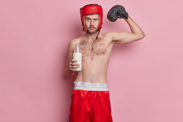 强壮严肃自信的运动员举起手臂展示他的强壮肌肉喝牛奶保持蛋白质饮食戴帽子和短裤赤裸上身站立白种人健身健美