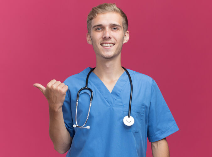 人面带微笑的年轻男医生穿着医生制服 听诊器指向隔离在粉红色墙上的一侧 并留有复印空间年轻人公民医生