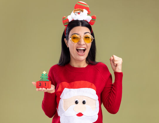 年轻快乐的年轻白人女孩戴着圣诞老人的头带和毛衣戴着眼镜拿着圣诞树玩具和日期看着相机眨眼做是的手势孤立在橄榄绿的背景上圣诞树日期眨眼