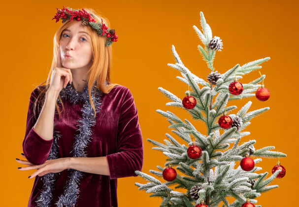 红色令人印象深刻的噘嘴年轻漂亮的女孩站在圣诞树附近穿着红色的裙子和花环与花环在脖子上把手指放在脸颊上孤立的橙色背景嘴唇花环手指