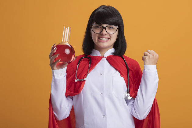 医学微笑着的年轻超级英雄女孩戴着听诊器 穿着医用长袍和斗篷 戴着眼镜 手里拿着装满红色液体的化学玻璃瓶 表示同意女性长袍液体