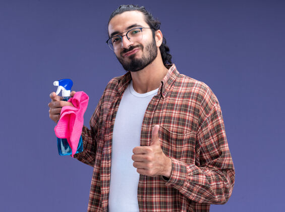 感觉高兴的年轻帅气的清洁工穿着t恤拿着喷雾瓶和抹布显示拇指向上隔离在蓝色的墙上男人年轻人