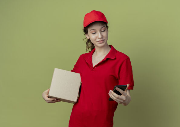 请很高兴年轻漂亮的送货女孩穿着红色制服 戴着帽子 拿着纸箱和手机 看着橄榄绿背景上的手机 还有复印空间橄榄纸箱帽子