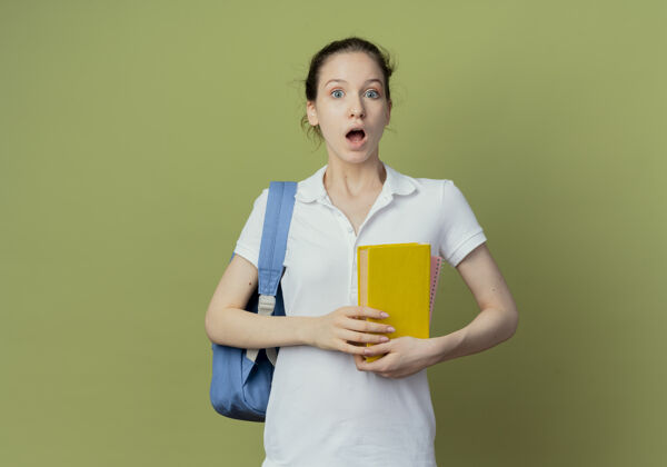 惊喜惊讶的年轻漂亮的女学生背着书包拿着书看着隔离在绿色背景上的相机和复印空间背绿色年轻