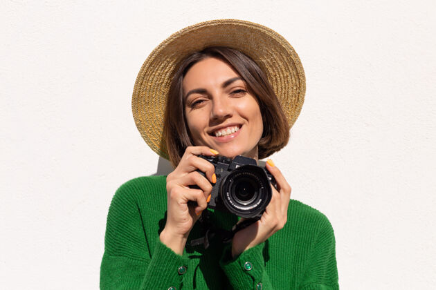 专业穿着绿色休闲毛衣和帽子的女士户外白墙上快乐的正面游客带着专业相机肖像墙年轻
