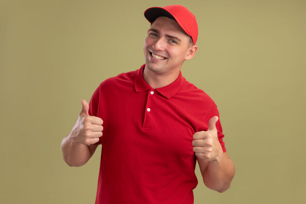 姿势微笑的年轻送货员穿着制服 戴着帽子 竖起大拇指 孤零零地站在橄榄绿的墙上脸帽子制服
