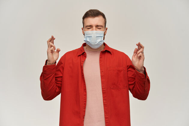 流感身穿红衫 脸上戴着防病毒面具 眼睛紧闭 手指交叉在白墙上许愿流行病掩护射击