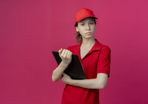 红色严格年轻漂亮的送货女孩穿着红色制服 戴着帽子拿着剪贴板 看着被隔离在深红色背景上的相机 还有复印空间背景举行剪贴板