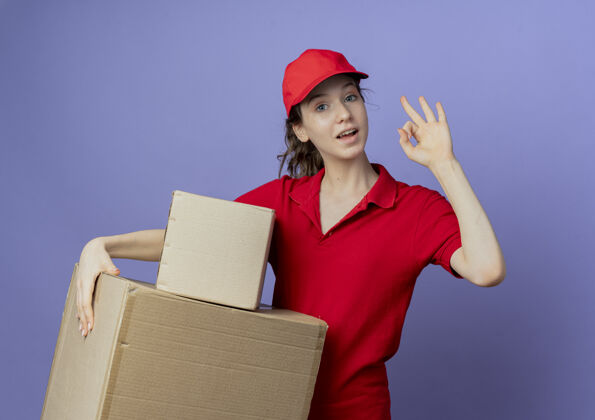 年轻令人印象深刻的年轻漂亮的送货女孩穿着红色制服和帽子举行纸箱和做ok标志孤立的紫色背景交货女孩好