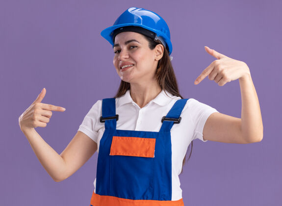 脸身着制服 面带微笑的年轻女建筑工人指着隔离在紫色墙上的自己表情人微笑