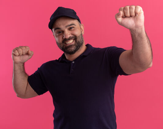表情微笑的中年送货员穿着制服 举起拳头 孤立地站在粉红色的墙上人人帽子