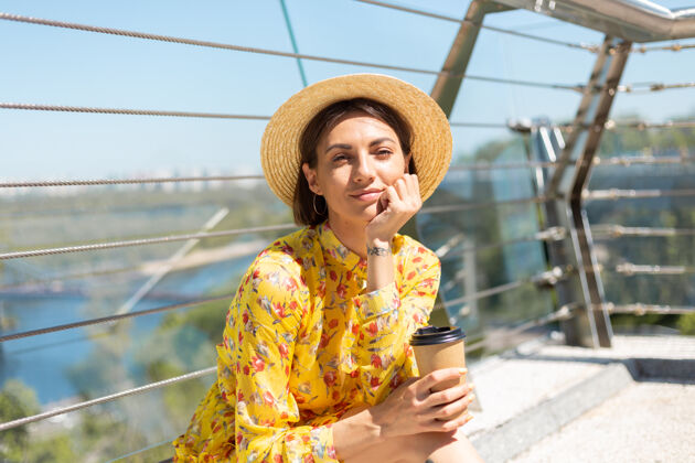 城市穿着黄色夏装 戴着帽子 喝着咖啡 享受阳光的女人的户外肖像户外请饮酒