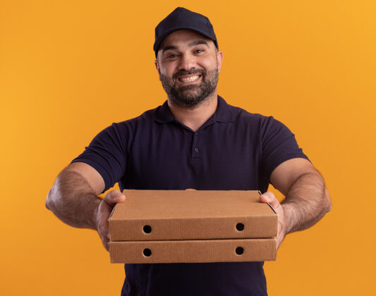 表情身着制服 头戴鸭舌帽 面带微笑的中年送货员把披萨盒放在隔离在黄色墙上的门前站着脸人