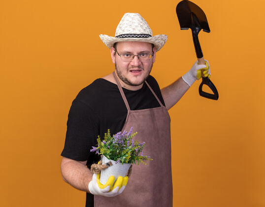 穿着高兴的年轻男园丁戴着园艺帽和手套 手里拿着花盆里的花 双手分开放在橙色的墙上园艺人站