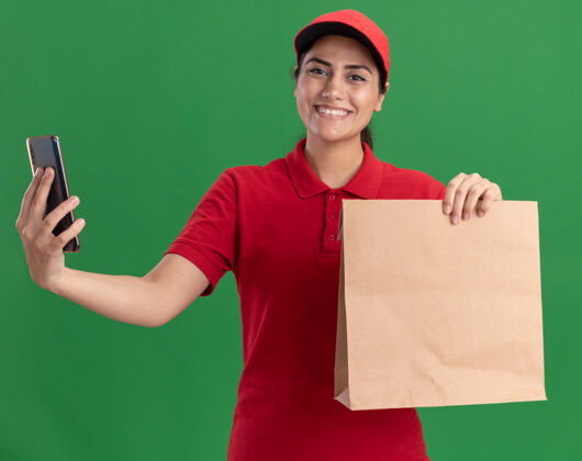 纸微笑着的年轻送货女孩穿着制服 戴着帽子 手里拿着纸食品包 电话被隔离在绿色的墙上站着女孩制服