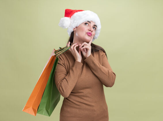 圣诞快乐令人印象深刻的仰望年轻漂亮的女孩戴着圣诞帽 肩上拿着礼品袋 橄榄绿背景上孤立着女孩向上包