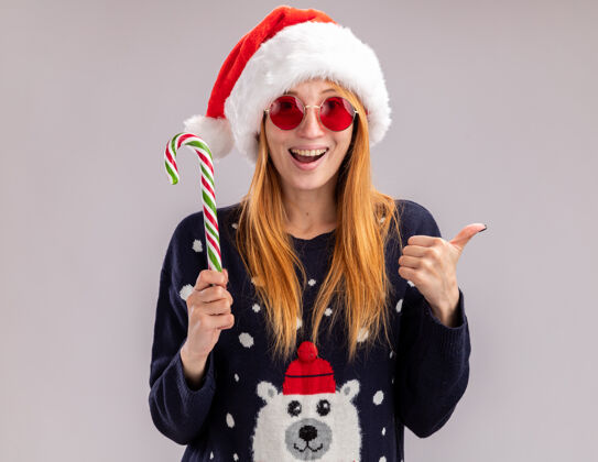 年轻快乐的年轻漂亮女孩戴着圣诞帽戴着圣诞眼镜拿着圣诞糖果在白色背景上孤立地竖起大拇指帽子女孩举行