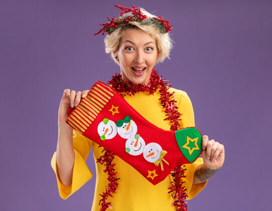 紫色兴奋的金发女郎头戴圣诞花环 脖子上戴着金属丝花环 手里拿着圣诞长袜 看着紫色背景上孤立的摄像机金发拿着花环