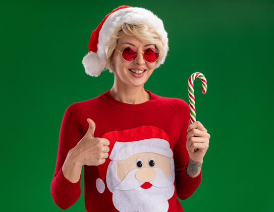 金发快乐的金发女郎戴着圣诞帽和圣诞老人的圣诞毛衣戴着眼镜看着相机拿着圣诞糖果手杖在绿色背景上孤立地竖起大拇指女人毛衣圣诞老人