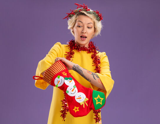 脖子好奇的金发女郎头戴圣诞花环 脖子上戴着金箔花环 手里拿着圣诞长袜 把手放进袜子里 看着紫色背景上孤立的袜子周围好奇紫色