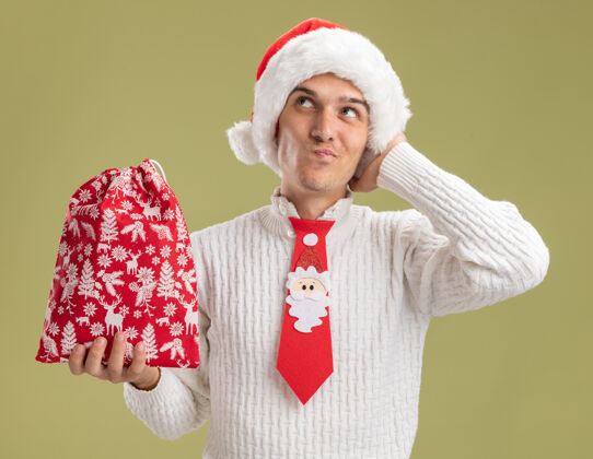 袋子可疑的年轻帅哥戴着圣诞帽 打着圣诞老人的领带 手里拿着圣诞袋 手放在头上 看着橄榄绿背景上孤立的一面壁板小伙子怀疑