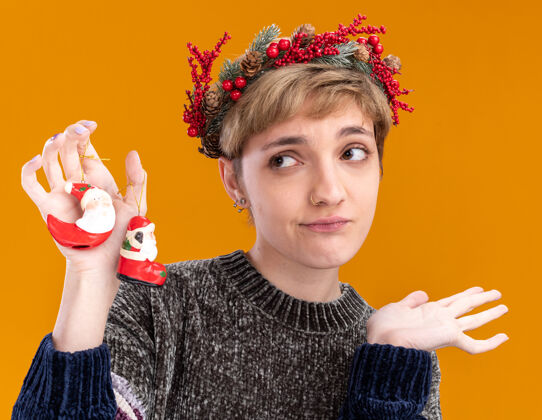 头不知所措的年轻漂亮女孩戴着圣诞花环手持圣诞老人的圣诞饰品望着一旁展示着孤立在橙色背景上的空手圣诞快乐装饰品壁板