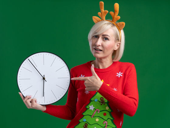 女人印象深刻的中年金发女人穿着圣诞驯鹿鹿角头带和圣诞毛衣拿着和指着时钟看着相机隔离在绿色背景圣诞节圣诞快乐金发