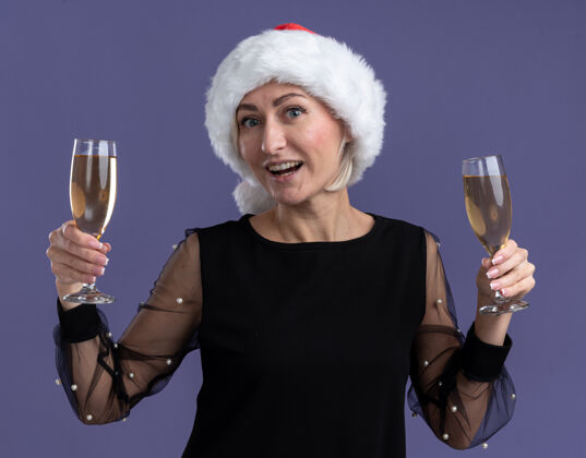 圣诞节快乐的中年金发女人戴着圣诞帽看着相机拿着两杯香槟隔离在紫色背景上金发圣诞快乐新年