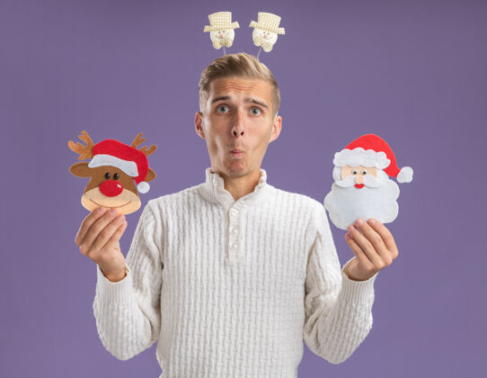 纸令人印象深刻的年轻帅哥戴着雪人头带拿着圣诞驯鹿和圣诞老人的纸饰品看着相机 噘起的嘴唇孤立在紫色的背景上装饰品圣诞老人小伙子
