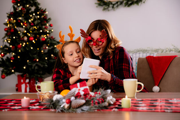 时间妈妈戴着驯鹿眼镜看着笔记本 女儿坐在桌旁 在家里享受圣诞节的时光桌子享受女儿