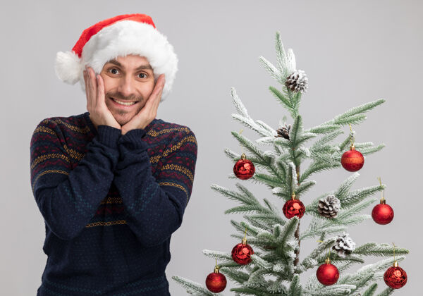 手兴奋的白人年轻人戴着圣诞帽站在圣诞树旁 手放在脸上看着隔离在白色背景下的摄像机圣诞快乐白色男人