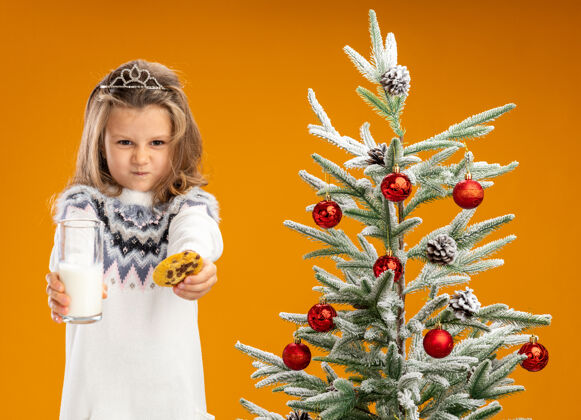 脖子皱着眉头的小女孩站在圣诞树旁 戴着头饰 脖子上戴着花环 手里拿着一杯牛奶和饼干 镜头被隔离在橙色背景下手拿小架子