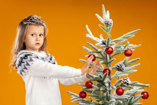 圣诞站在圣诞树旁 戴着头饰 脖子上戴着花环的小女孩心事重重地把手伸向孤立在橙色背景下的圣诞树花环关心站