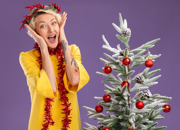 圣诞树兴奋的金发女郎头戴圣诞花环 脖子上戴着金属丝花环 站在装饰好的圣诞树旁 手放在脸上 看着紫色背景上孤立的摄像机女人手紫色