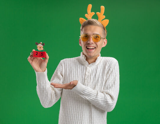 印象印象深刻的年轻帅哥戴着驯鹿鹿角头带戴着眼镜拿着圣诞树玩具和日期指着它用手看着相机隔离在绿色的背景圣诞手指向