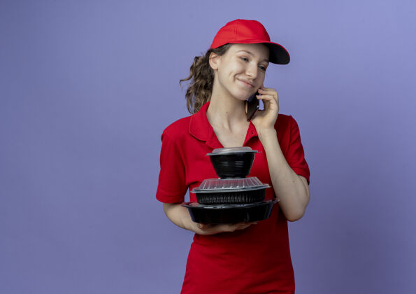 制服高兴的年轻漂亮的送货女孩穿着红色制服 戴着帽子 看着一边拿着食物容器 一边在紫色背景上讲着电话 还有复印空间请漂亮背景