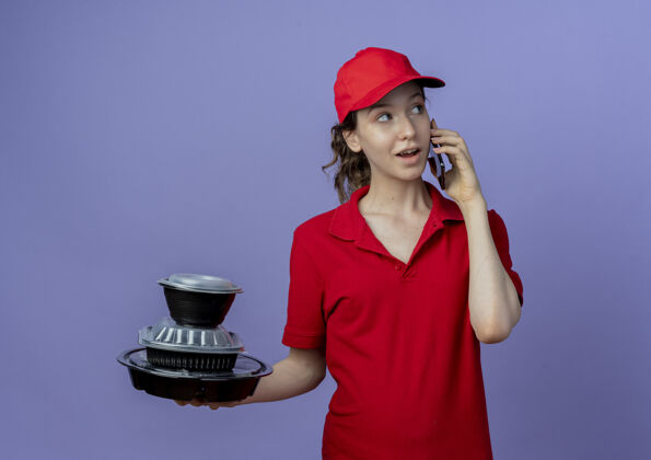 印象令人印象深刻的年轻漂亮的送货女孩穿着红色制服 戴着帽子 看着一边拿着食品容器 一边在紫色背景下讲电话年轻紫色电话