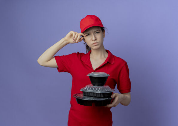 漂亮困惑的年轻漂亮的送货女孩穿着红色制服 戴着帽子拿着食物容器 眼睛被隔离在紫色背景上 还有复印空间容器眼睛举行