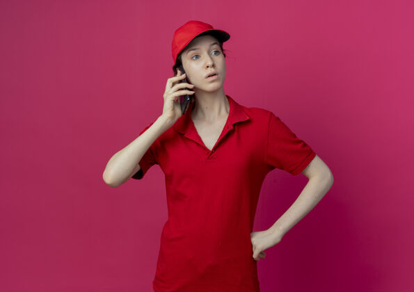 印象令人印象深刻的年轻漂亮的送货女孩穿着红色制服 戴着帽子打电话 手放在腰上 在深红色的背景上孤立地看着一边 还有复印空间背景漂亮制服