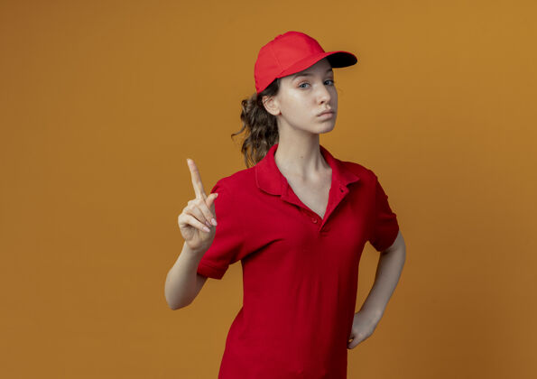 帽子自信的年轻漂亮的送货女孩 穿着红色制服 戴着帽子 手放在腰上 手指举在橙色背景上 留有复印空间信心制服手指