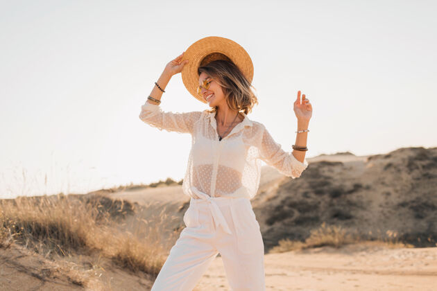 休闲落日时分 穿着白色衣服 戴着草帽和太阳镜 在沙漠沙滩上摆出一副时髦 快乐 迷人 微笑的女人造型时尚人模特