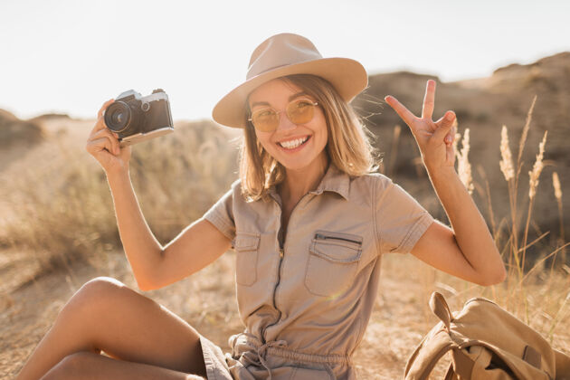 体验沙漠中穿着卡其色长裙的迷人时尚年轻女子 在非洲旅行 戴着帽子和背包 用老式相机拍照旅行相机配件