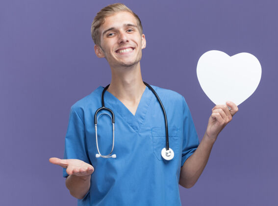 姿势微笑的年轻男医生穿着医生制服手持听诊器手持心形盒子指着隔离在蓝色墙上的摄像头人年轻人形状