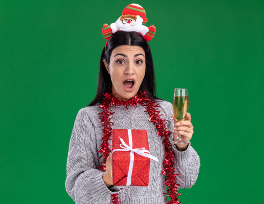 香槟年轻的白人女孩戴着圣诞老人的头带 脖子上戴着金箔花环 手里拿着礼包和一杯香槟 看着隔离在绿色背景上的相机印象复制玻璃
